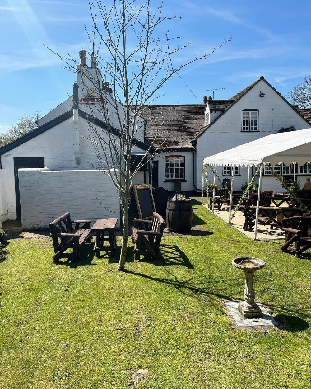 The garden at The Holly Bush: a wide lawn with heavy wooden benches, a stone bird bath and a white marquee along the side of the whitewashed pub.